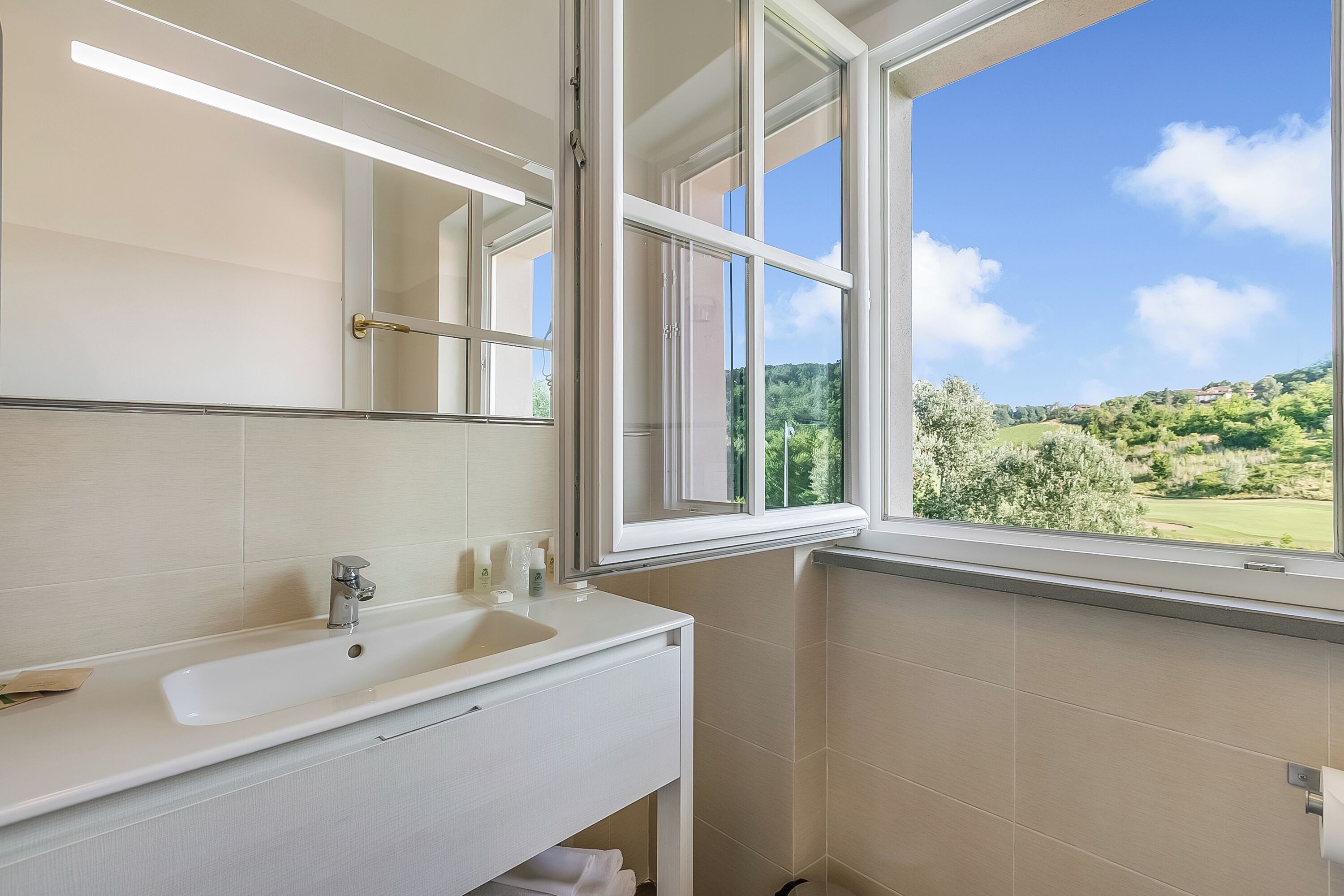 Un bany amb un lavabo blanc, un mirall i una finestra que mostra un paisatge verd.