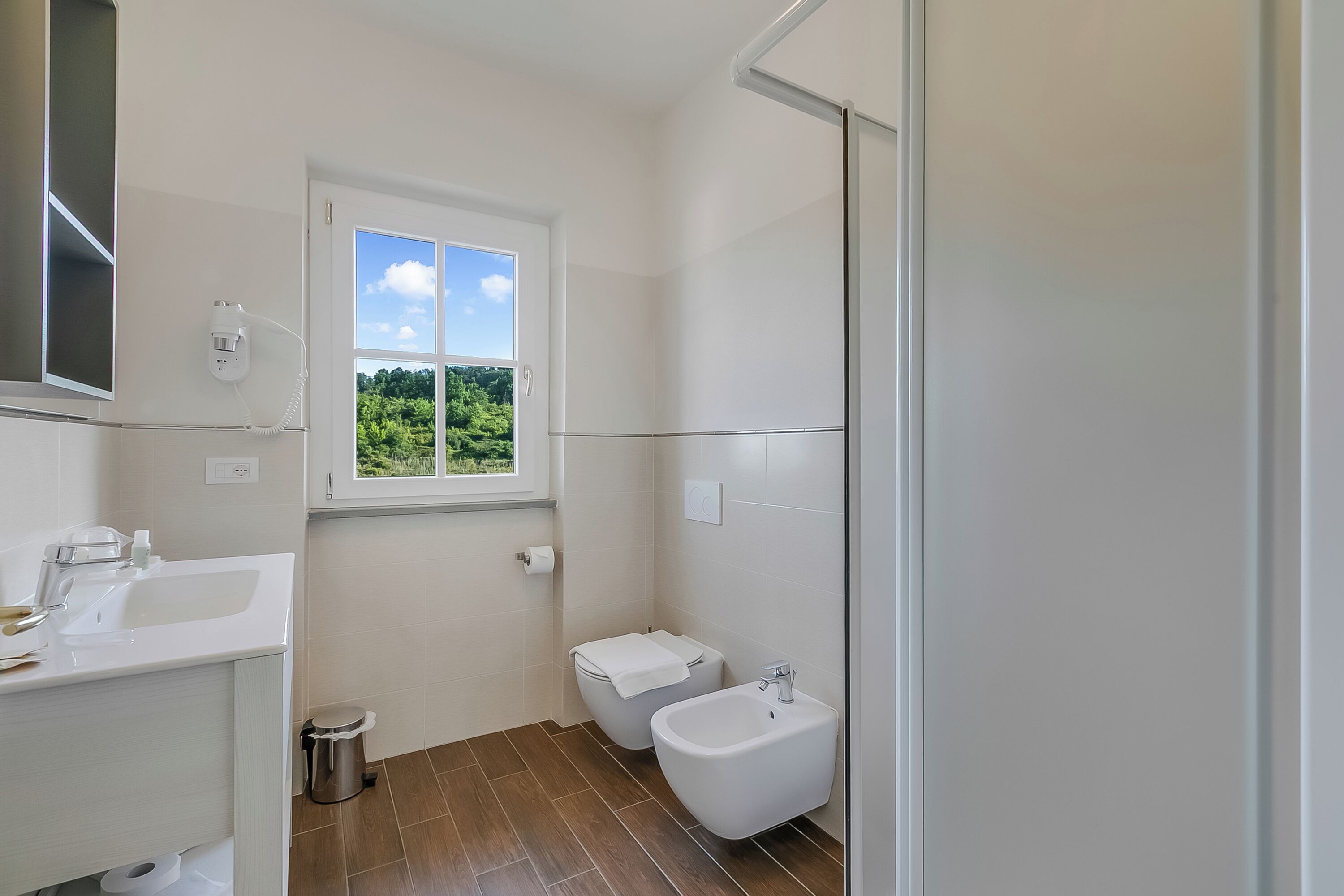 Ein Badezimmer mit Toilette, Bidet, Waschbecken und einem Fenster mit Blick ins Grüne.