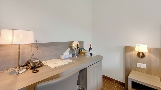 Habitación de hotel con un escritorio, un teléfono, una lámpara, un espejo, una botella de vino y una pequeña mesa con vasos.