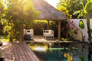 Un espace piscine doté d’une terrasse en bois, de deux chaises longues blanches, d’une structure à toit de chaume et de plantes tropicales.