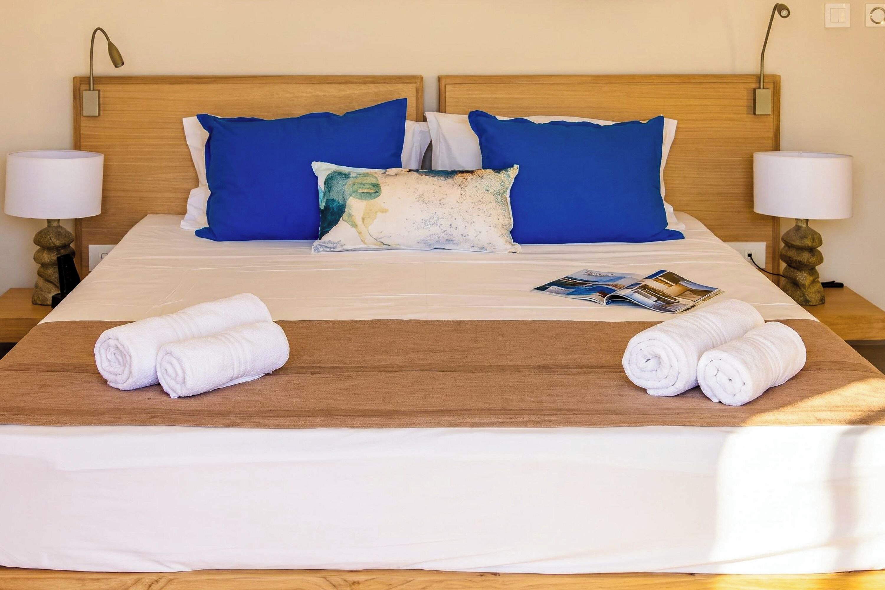 Una cama bien tendida con almohadas azules y blancas, un corredor marrón y dos toallas enrolladas sobre una plataforma de madera.