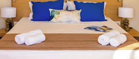A neatly made bed with blue and white pillows, a brown bed runner, and two rolled towels on a wooden platform.