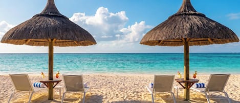 A beach with two thatched umbrellas and lounge chairs, each with a drink.