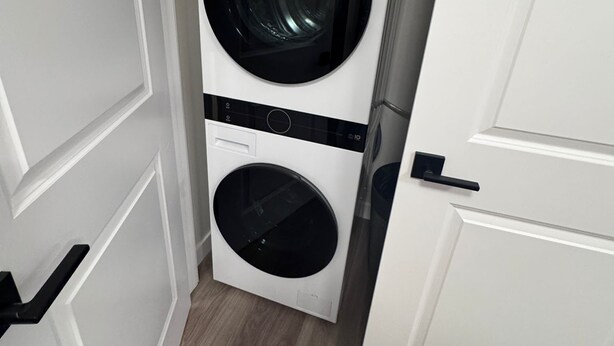 A white laundry room with two stacked washing machines, a door handle, and a wooden floor. - Serenity Suite (Regina)