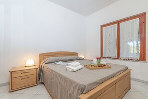 Ein ordentlich bezogenes Bett mit Holz-Kopfteil, ein Nachttisch mit Lampe und ein Fenster mit Vorhängen.