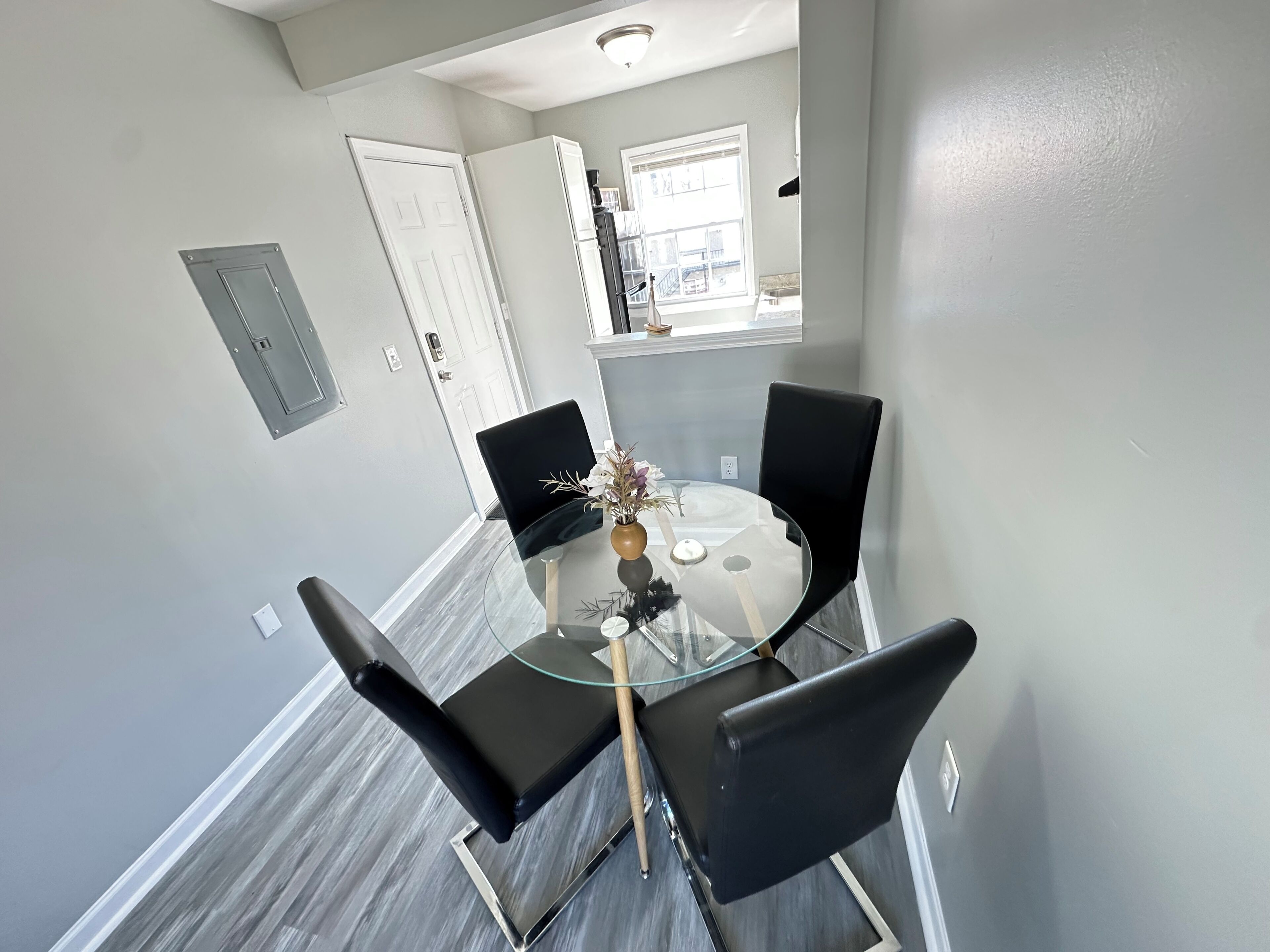 A dining area with a glass table and four black chairs, a white door, and a window with a view.