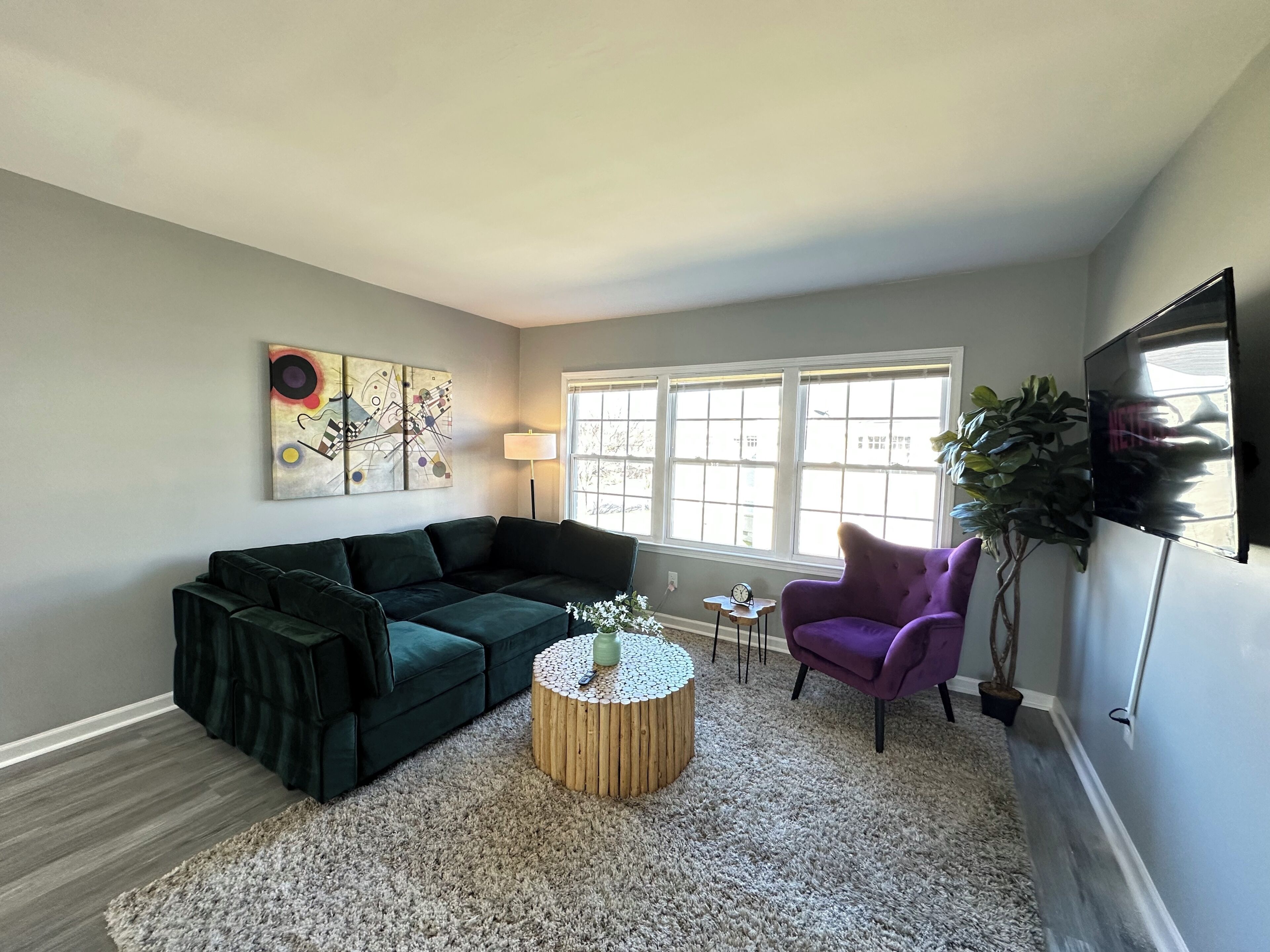 A living room with a dark green sofa, a round wooden coffee table, a purple armchair, and a large window with natural light.