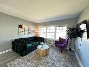 Un salon avec un canapé vert foncé, une table basse ronde en bois, un fauteuil violet et une grande fenêtre laissant entrer la lumière naturelle.