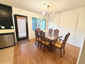 A dining area with a glass table and wooden chairs, a kitchen with a dishwasher, and a door with a window. - Southern charm awaits at Cobblestone Cottage (Wilmington)