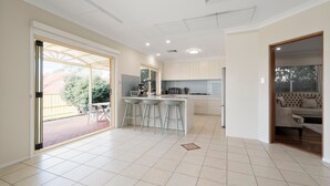 A modern kitchen with a central island, bar stools, and a dining area with a view of the garden. - Drumbern Pool House Macquarie Links (Macquarie Links)