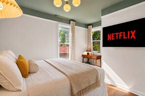 A bedroom with a bed, pillows, a bedside table, a lamp, a window with curtains, and a wall-mounted television displaying the Netflix logo. - Louisville Retreat | Hot Tub, Ping Pong, 3 Kings (Louisville)