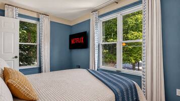 Une chambre à coucher avec un lit, un téléviseur affichant le logo « NETFLIX », et une fenêtre avec des rideaux.