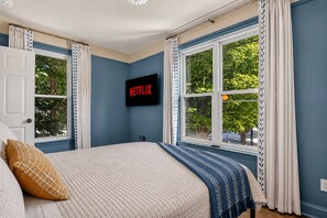 Ein Schlafzimmer mit einem Bett, einem Fernseher, der 'NETFLIX' anzeigt, und einem Fenster mit Vorhängen.
