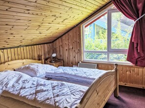Ein Zimmer mit Holzboden und schrägem Dach, ein Bett mit gesteppter Decke, ein Nachttisch mit Lampe sowie ein Fenster mit Vorhängen.