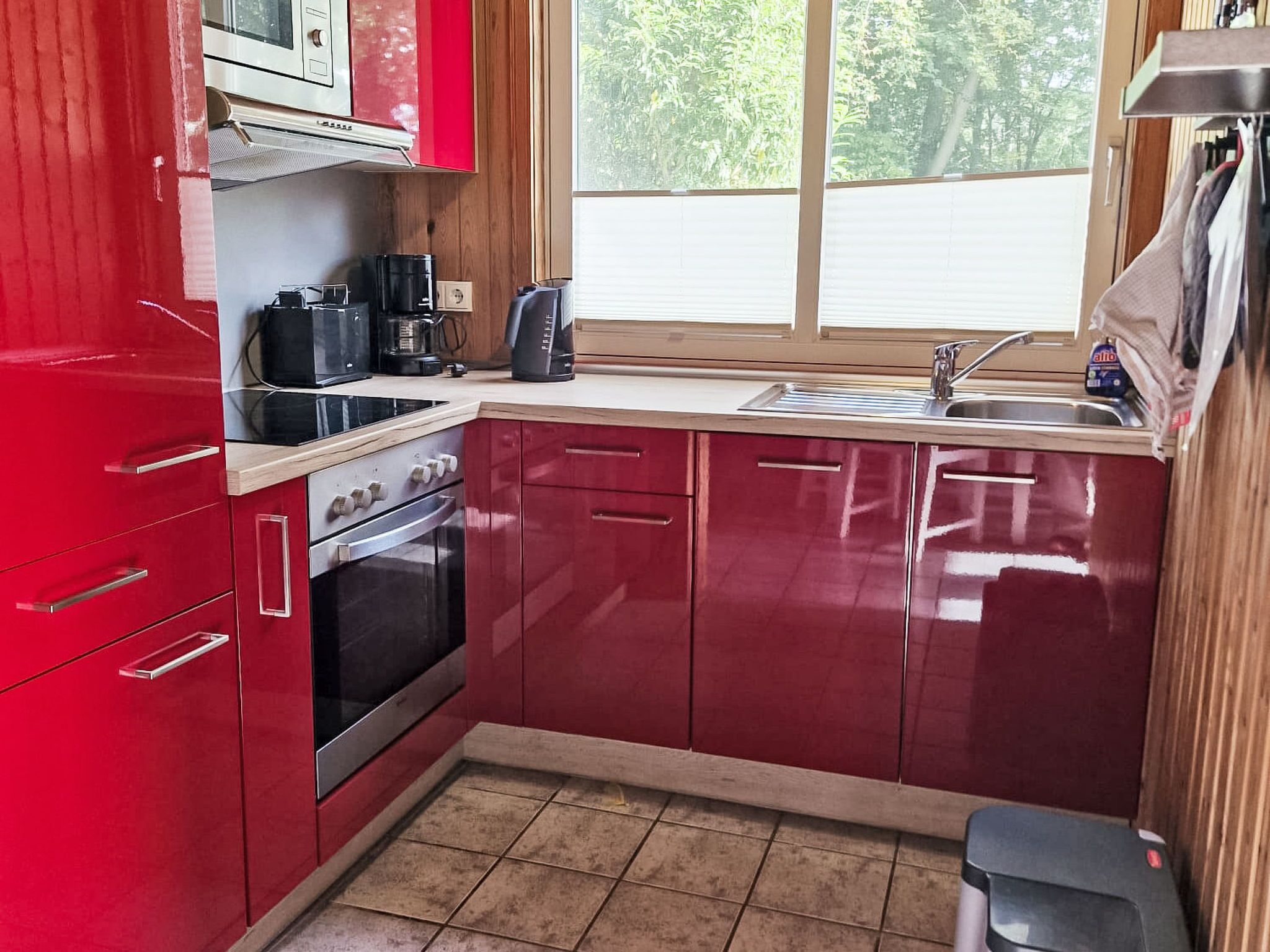 A compact kitchen with red cabinets, a stove, a sink, and a window with a view of greenery outside.