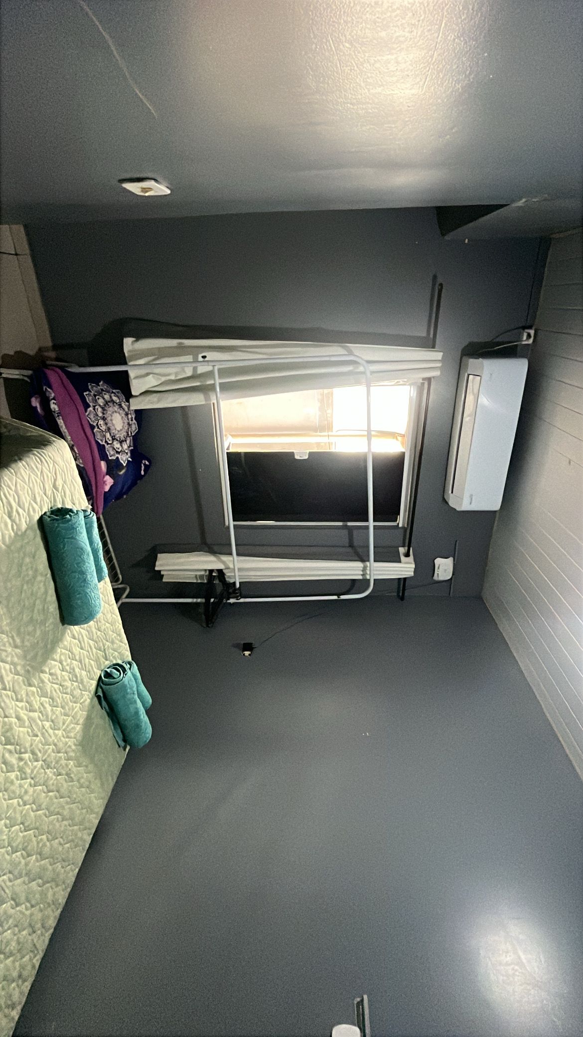 Una cama litera con cabecera y una mesita de noche pequeña, un calentador fijado a la pared, y una ventana con persianas.
