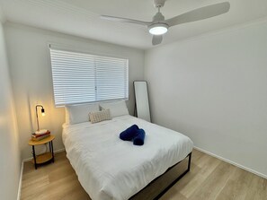Une chambre à coucher comprenant un lit, un ventilateur de plafond, une table de chevet avec une lampe et une fenêtre avec des stores.