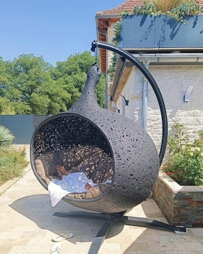 A metallic sculpture resembling a teapot with a person inside, placed outdoors on a paved area with a stone wall and greenery in the background. - Gîtes de France® - Logis de la Mésange Bleue (GIGNAC)