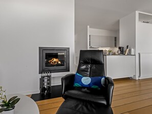 Ein modernes Wohnzimmer mit Kamin, einem schwarzen Ledersessel mit Kissen und einer Küchenzeile im Hintergrund.
