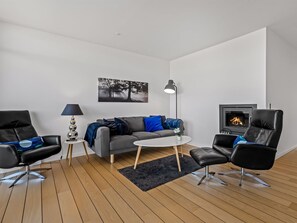 Un salon moderne avec un canapé gris, deux fauteuils en cuir noir, une table basse ronde, un tapis noir, un lampadaire, une cheminée et un tableau encadré accroché au mur.