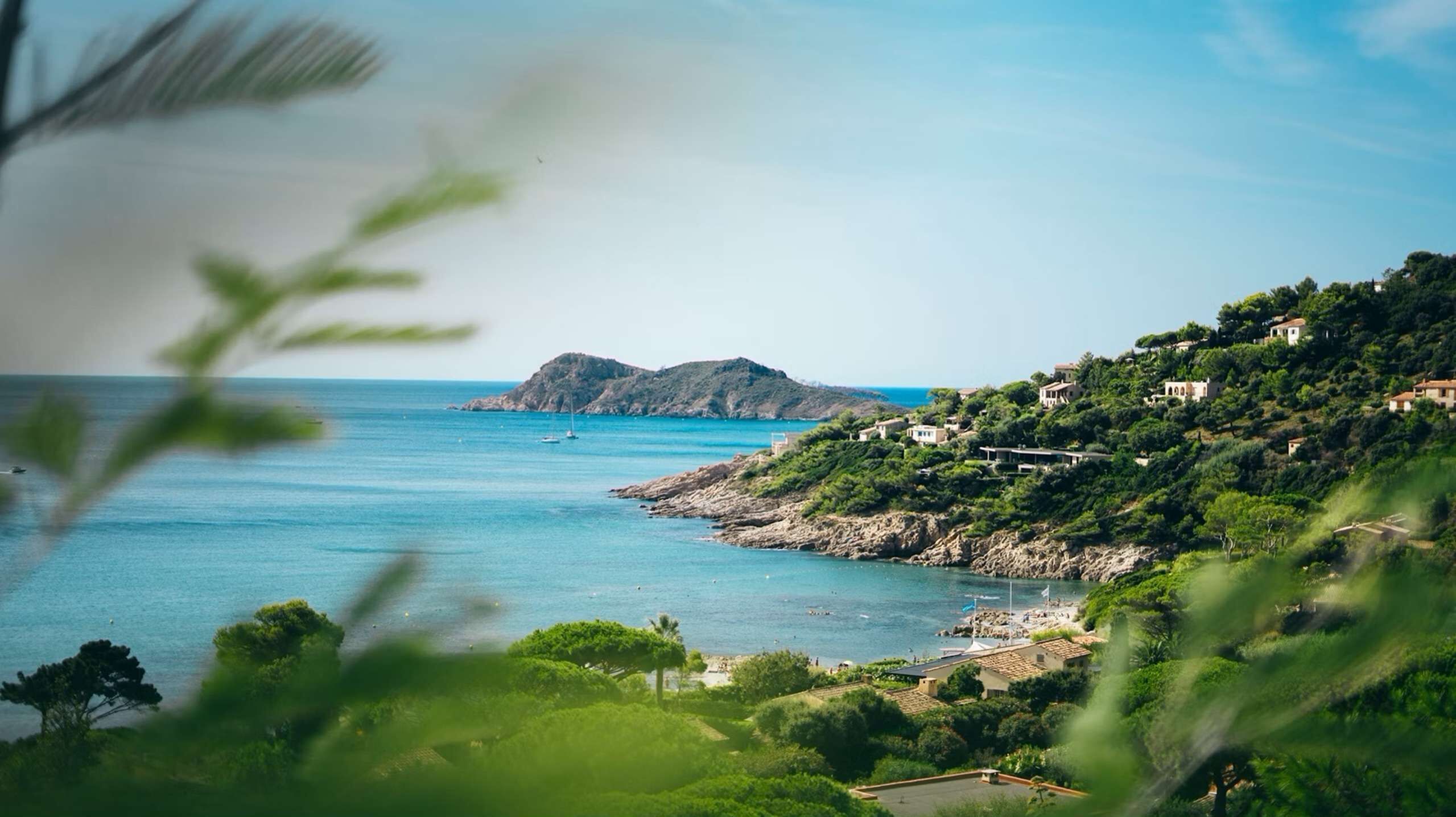 A coastal landscape with a rocky shoreline, lush greenery, and buildings on a hillside.