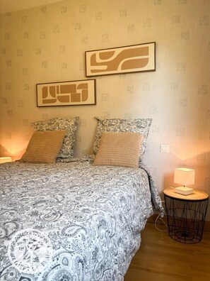 A bedroom with a bed, patterned bedding, two wall-mounted abstract art pieces, and a bedside lamp on a small table. - 46G13379 - PRENDEIGNES (PRENDEIGNES)