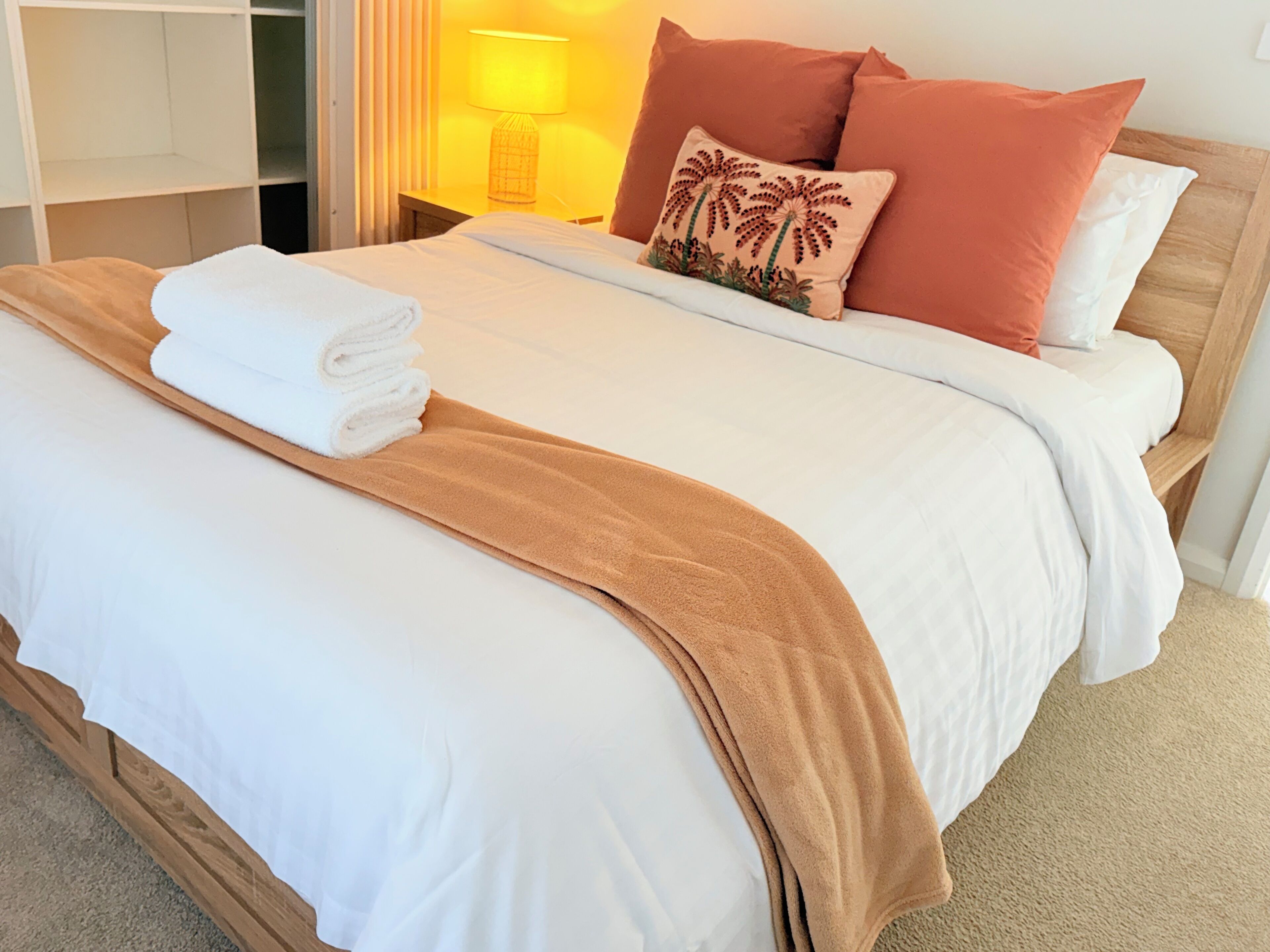 A neatly made bed with white and brown linens, a folded towel, and a single decorative pillow.