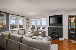 A living room with a sofa, chairs, a fireplace, a TV, and a view of the sea through large windows. - Ocean Views, Hot Tub, Fireplaces & Luxury Finishes, Chef's Kitchen, Community Pool, EV Charger, BBQ (Lincoln City)