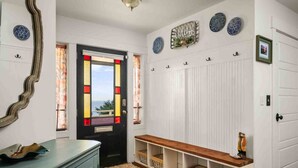 A hallway with a black door featuring stained glass, a white wall with decorative plates, and a bench with storage baskets. - Ocean Views, Hot Tub, Fireplaces & Luxury Finishes, Chef's Kitchen, Community Pool, EV Charger, BBQ (Lincoln City)