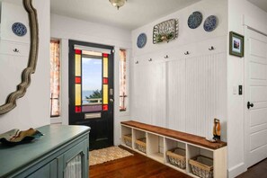 A hallway with a black door featuring stained glass, a white wall with decorative plates, and a bench with storage baskets. - Ocean Views, Hot Tub, Fireplaces & Luxury Finishes, Chef's Kitchen, Community Pool, EV Charger, BBQ (Lincoln City)