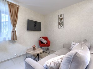 Ein modernes Wohnzimmer mit einer Couch, einem roten Sessel, einem kleinen Tisch mit einer Vase, einem Flachbildfernseher und einem dekorativen Blattbild an der Wand.