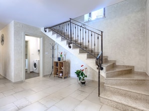 Eine Treppe mit Metallgeländer führt zu einer oberen Etage, daneben ein Garderobenhaken, ein Blumentopf und ein Waschraum mit einer Waschmaschine.