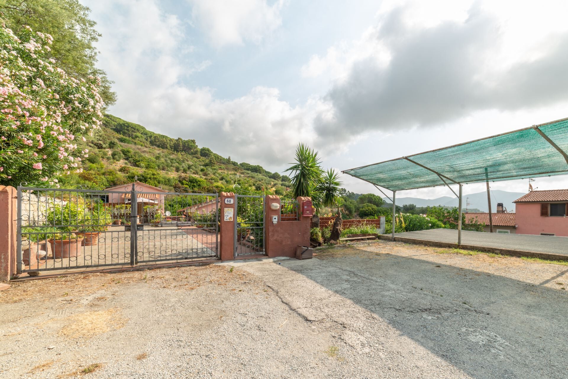 A gated entrance to a property with a gravel driveway, a covered area with a green roof, and a hillside with vegetation.