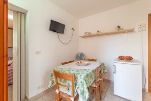 A compact dining area with a table, chairs, a television mounted on the wall, a small refrigerator, and a shelf with items. - APPARTAMENTI I LIMONI - APPARTAMENTI I LIMONI - BILO 4 (Campo nell'Elba)