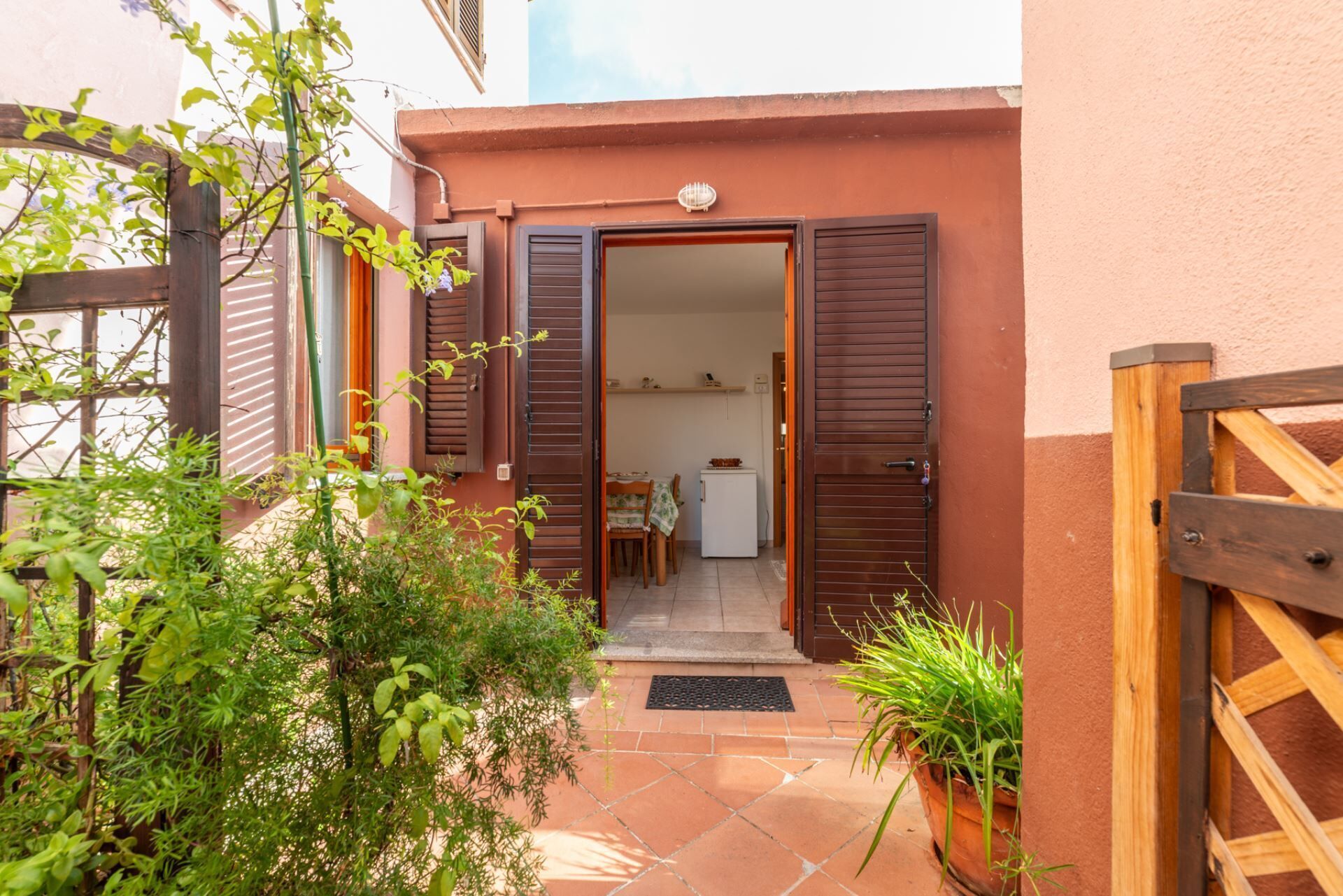 A residential entrance with a wooden gate, a tiled patio, and a small dining area visible through the open door.