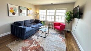 A living room with a blue sofa, a red chair, a coffee table, and a large window with blinds. - 1BR Business Suite with Parking Near Downtown Birmingham (Birmingham)
