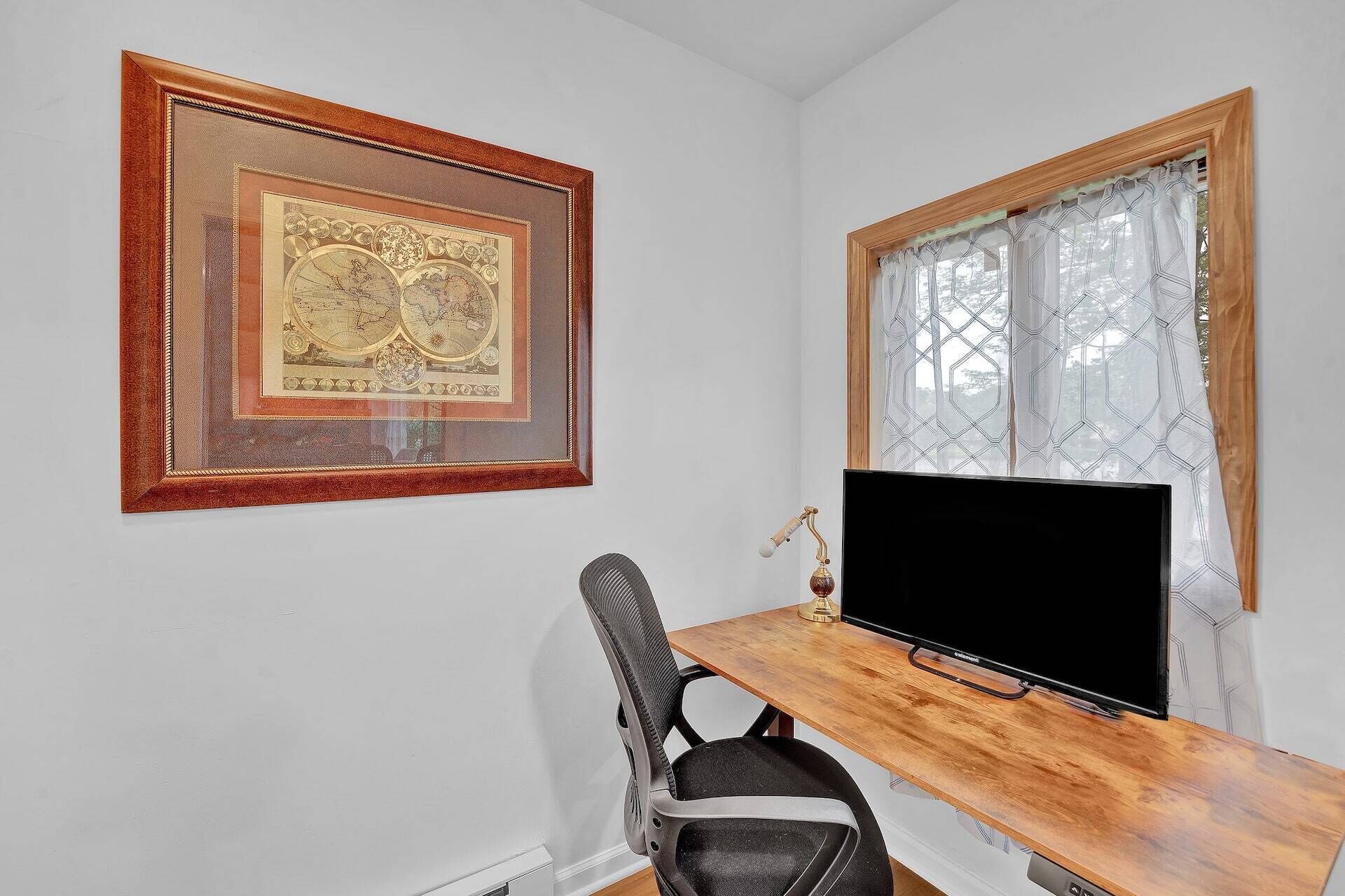 Una habitación con un escritorio de madera, una silla negra, un mapa enmarcado en la pared y una ventana con cortinas estampadas.