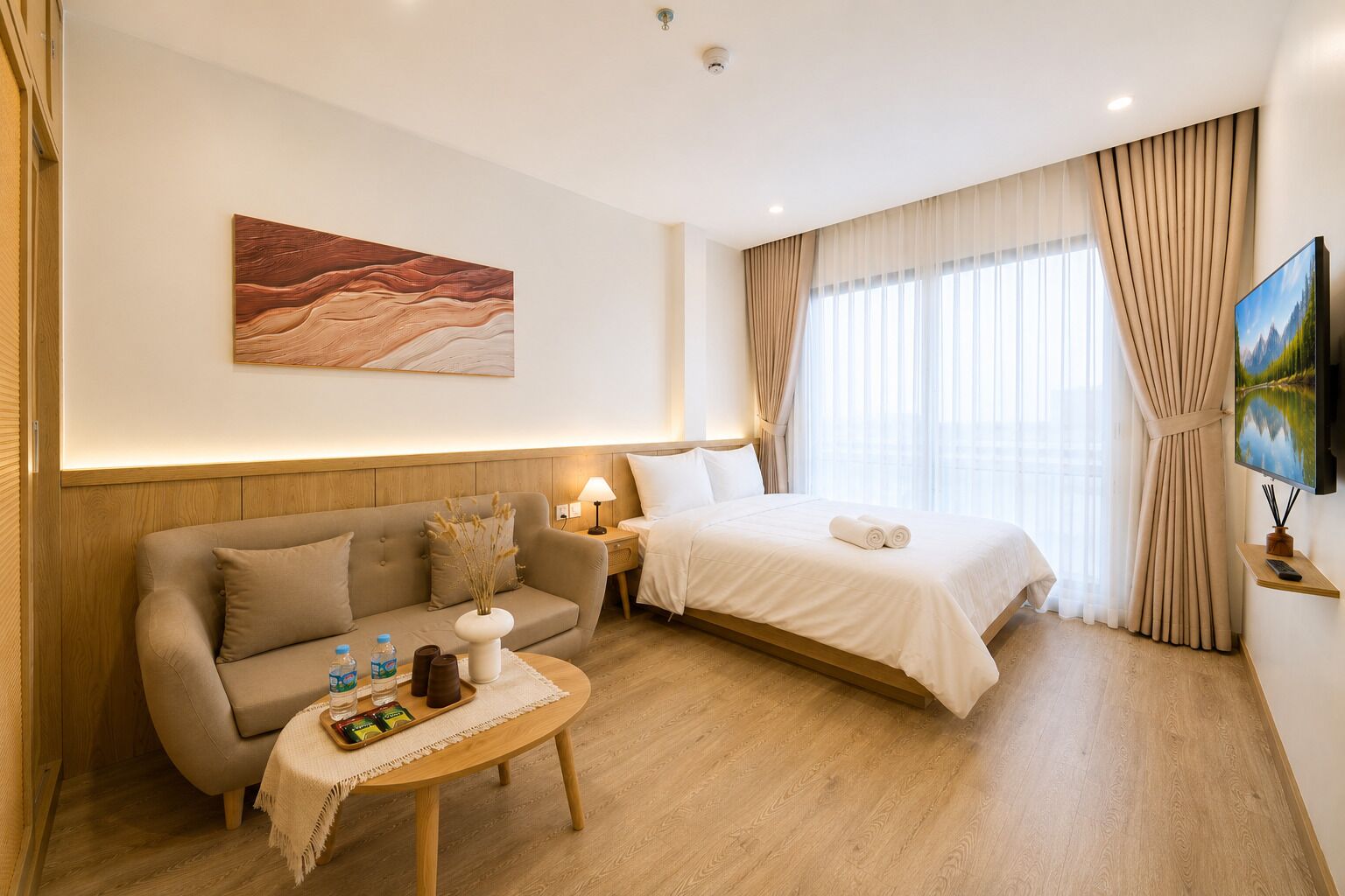 A hotel room with a bed, a sofa, a small table with a vase and bottles, a painting on the wall, and a TV on a stand.