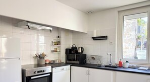 A modern kitchen with white cabinetry, a stainless steel range, a built-in oven, and a window with a view of a stone building outside. - Gîtes de France® - La Maison de la Rivière (GINTRAC)