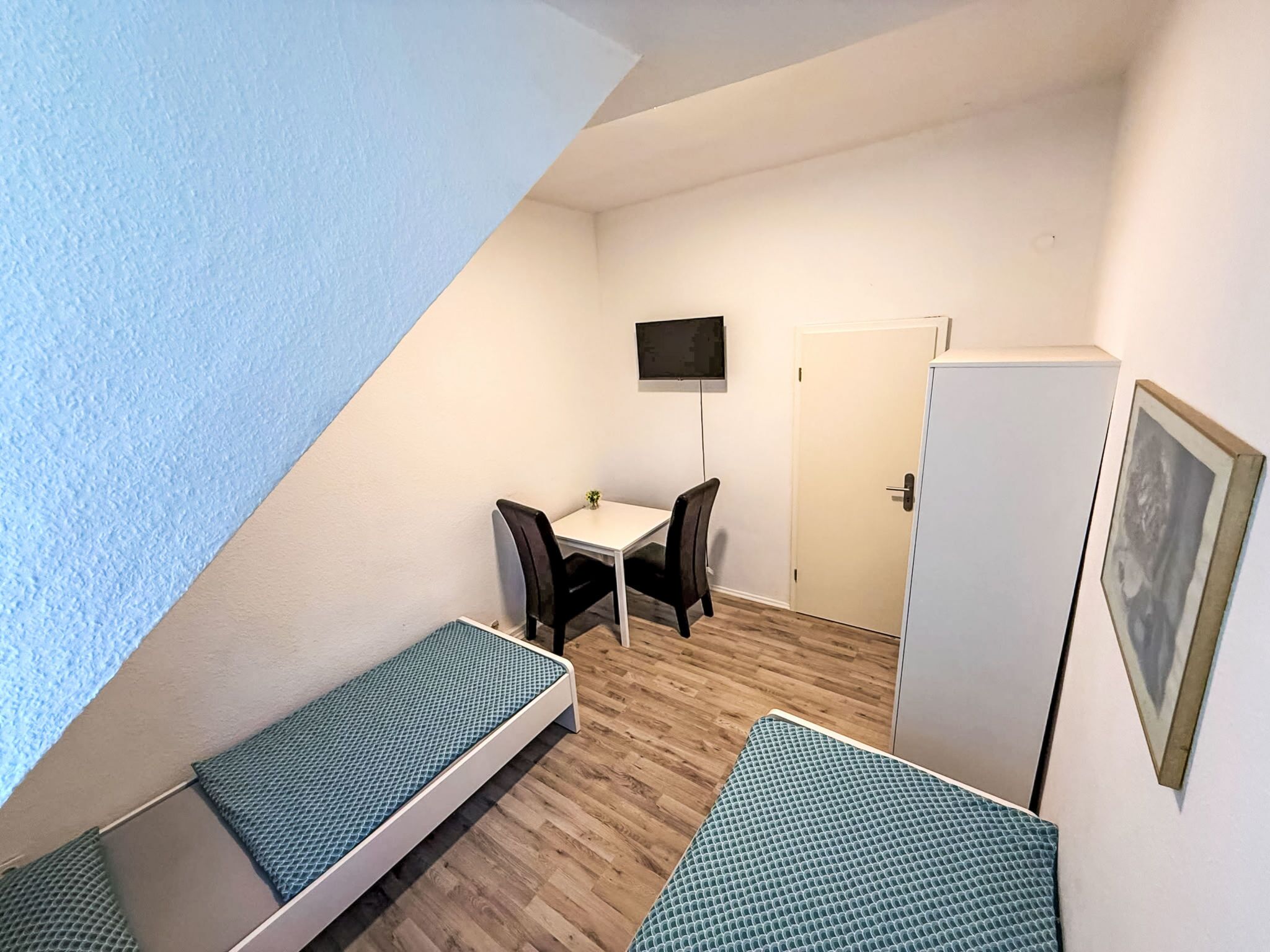 A small, triangular room with two single beds, a desk with a chair, a television mounted on the wall, and a door.