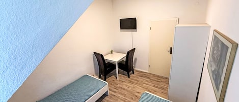 A small, triangular room with two single beds, a desk with a chair, a television mounted on the wall, and a door.