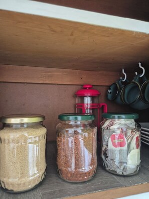 Une étagère avec trois bocaux en verre contenant divers aliments, une cafetière rouge et verte, et des pots suspendus.