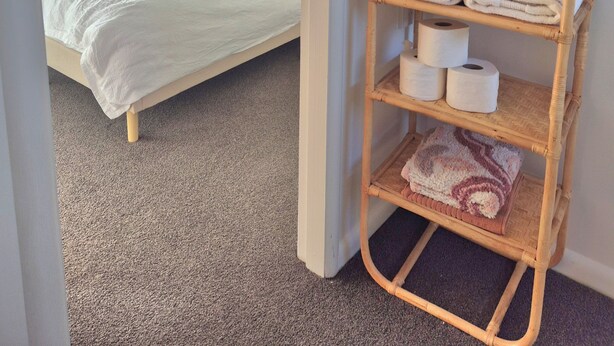 A bedroom with a bed, a bedside shelf with towels and toiletries, and a window with curtains. - Tāhunanui Nest (Nelson)