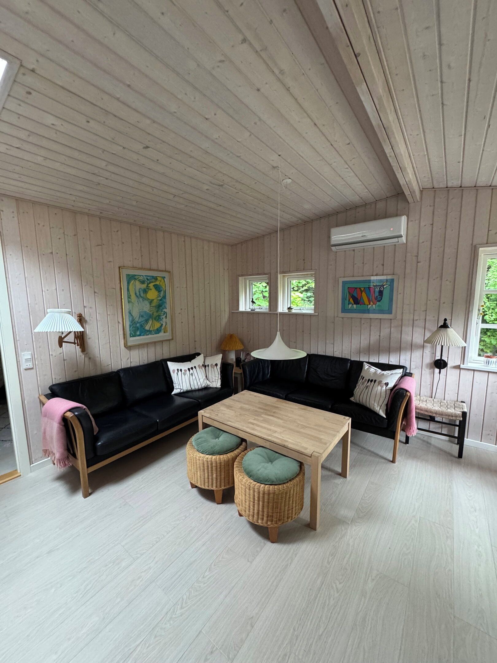 A room with a wooden ceiling, a black sofa with cushions, a wooden coffee table, wicker ottomans, and a lamp.