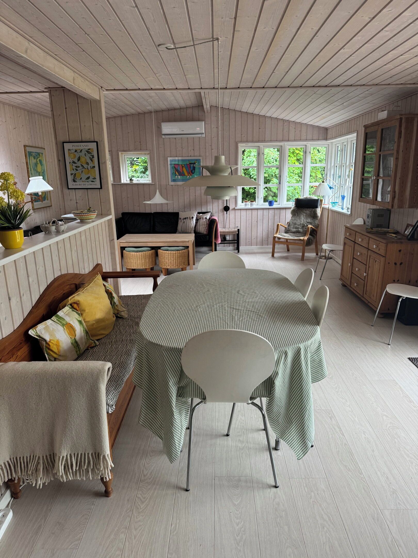 A spacious room with a round dining table, a sofa, and a chair. There are windows with views of greenery outside.