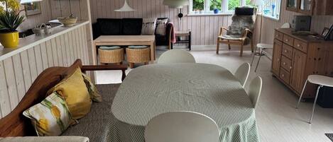 A spacious room with a round dining table, a sofa, and a chair. There are windows with views of greenery outside.