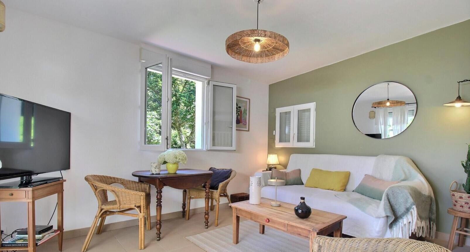 Un salon comprenant un téléviseur, une table à manger, des chaises, un canapé, une table basse, un miroir et une fenêtre donnant sur un paysage verdoyant.