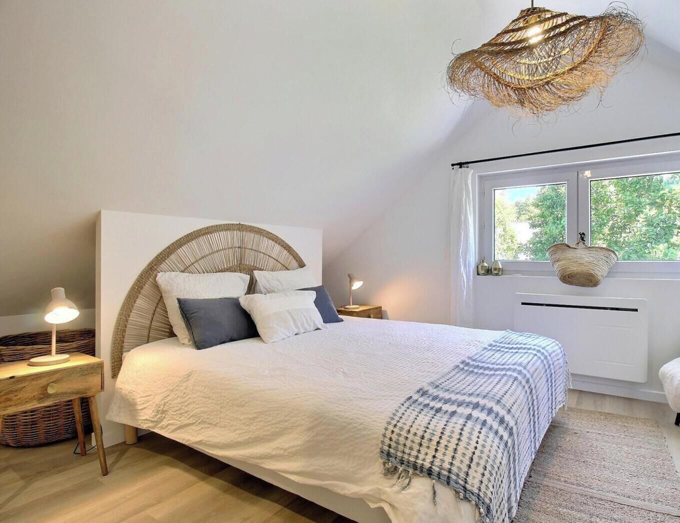 Ein Schlafzimmer mit einem Bett, Nachttischen, einer Hängelampe, einem Fenster mit Blick und einem Korbbrettchen am Fensterbrett.