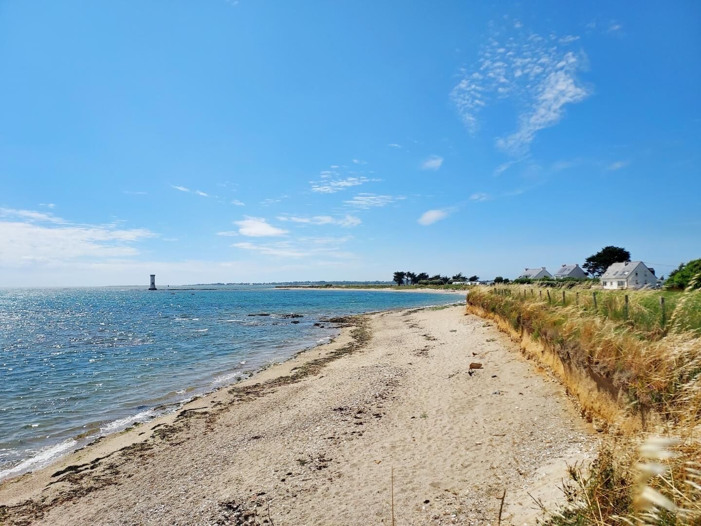 A beach with a clear sky, a lighthouse in the distance, and houses along the shore.