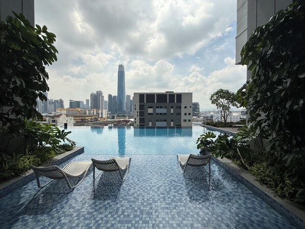 Une piscine à débordement située sur le toit, avec des chaises longues, entourée de verdure et offrant une vue imprenable sur les gratte-ciel d’une ville moderne.